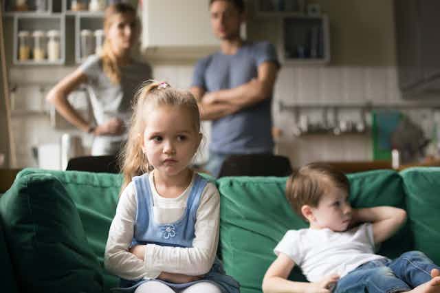 Niña enfadada sentada junto a su hermano en el sofá mientras sus padres los observan desde el fondo.