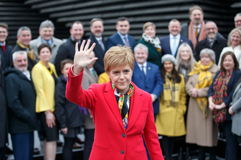 Nicola Sturgeon waving bit looking serious in front of her SNP colleagues.