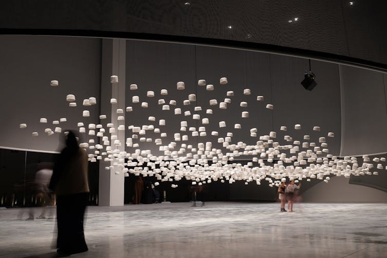 Figures stand underneath a sculptural installation made up of hundreds of hats, glowing white in a dark room.
