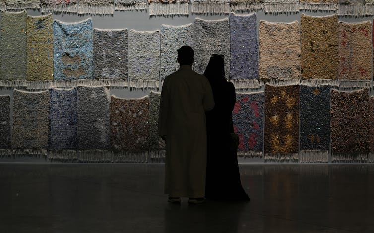 A couple stand in silhouette viewing a tapestry of work that is constructed from prayer rugs of different colours and textures.