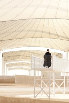 A woman in a black dress stands on a platform in a vast interior space.