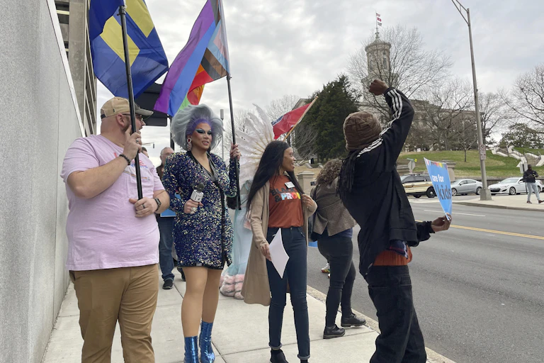 Protesters carrying banners walk along a sidewalk with a state building in the background.