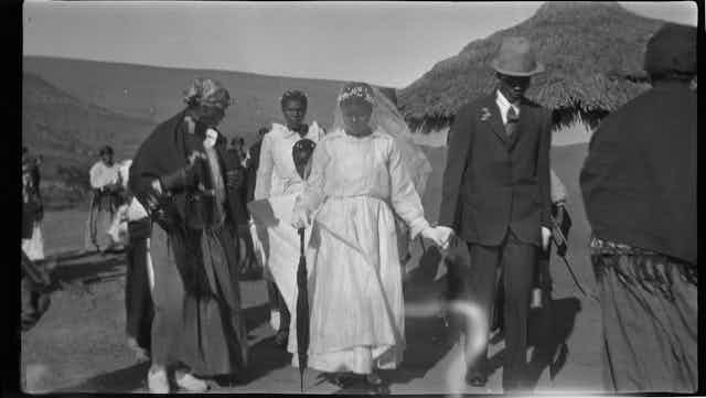 An old black-and-white photo shows a traditional rural setting, where an African couple walks hand-in-hand in formal western wedding attire, past villagers in more traditional attire.