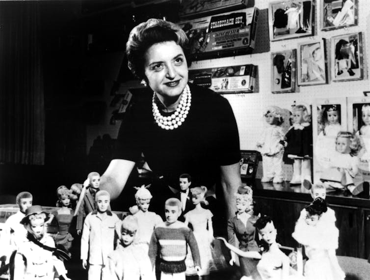 Black and white photo of Ruth Handler, Barbie inventor, standing behind a display of male and female dolls.