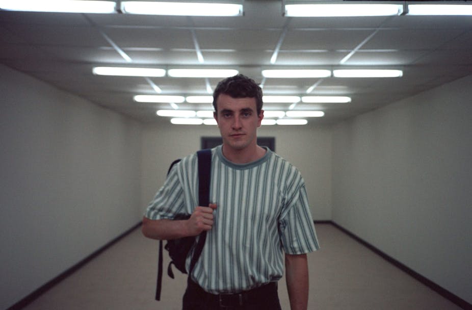 Paul Mescal stands in a bare corridor with a rucksack over one arm, staring straight into camera.