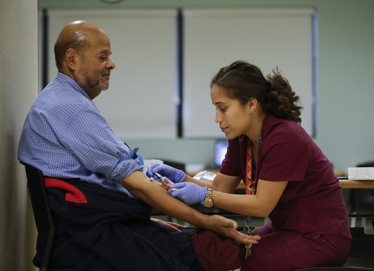 A medical technician takes a blood sample from a man sitting in a chair.