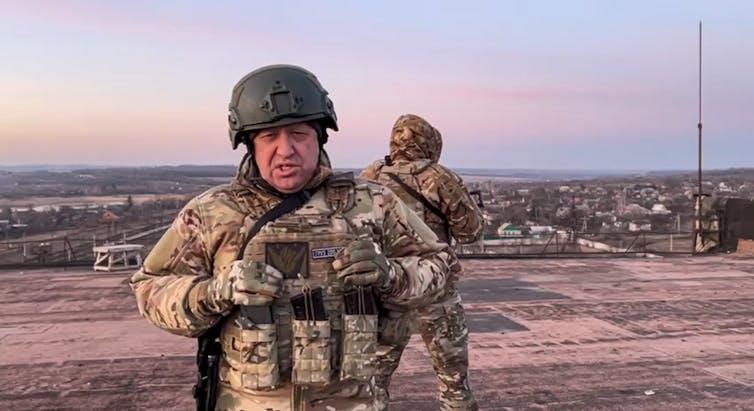 An older man in battle fatigues and a helmet looks into a camera with another soldier and a pink sky behind him.