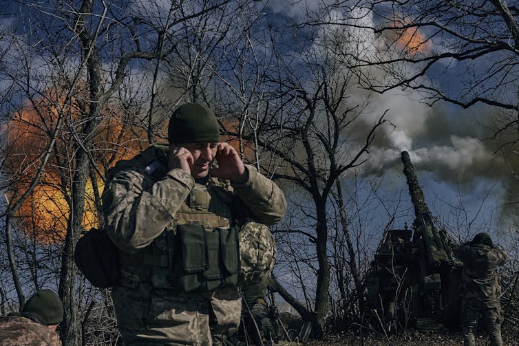 A soldier holds his hands to his ears as large puffs of fire and smoke are seen through the branches of trees behind him.
