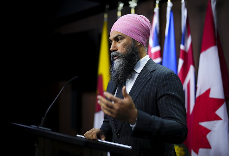 An Indian man in a pink turban and a suit speaks at a podium in front of a Canadian flag