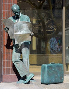 A statue of a man leaning against a building and reading a newspaper