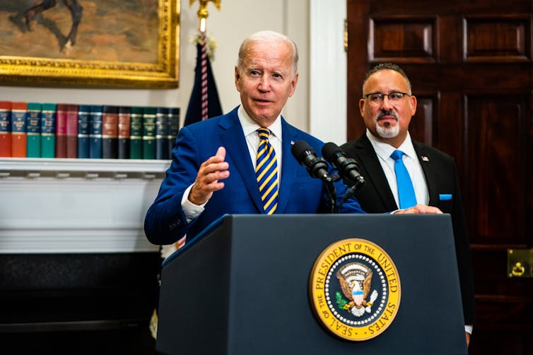 A gray haired man in a royal blue suit jacket with a royal blue and yellow tie, speaks from behind a lectern. A gray bearded, bespectacled man, wearing a black suit and cobalt blue tie, stands behind him and to the left.