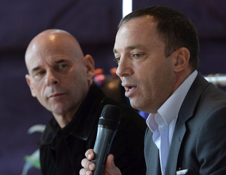 A man speaks into a microphone while another man, who is bald, looks on