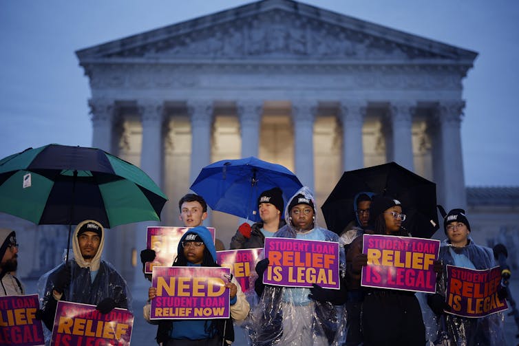 A group of nine young men and women stand in the evening rain, some hold umbrellas. Others wear hats or hoodies. Each of them holds a purple, pink, orange and yellow sign that reads: Student loan relief is legal.