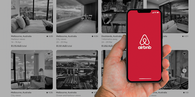 A hand holding a phone with Airbnb logo, with search results for available accomodation displayed in the background. 