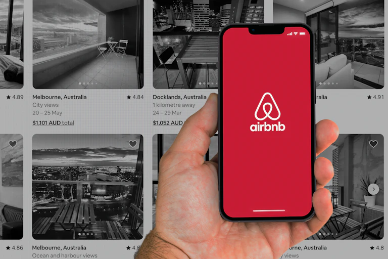 A hand holding a phone with Airbnb logo, with search results for available accomodation displayed in the background.