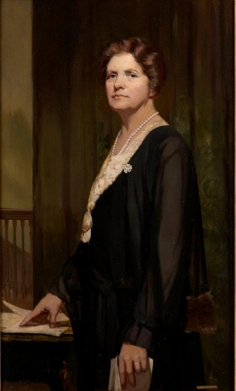 A portrait of a white woman with brown wavy hair. She is wearing a white blouse with pearls and a see through cardigan.