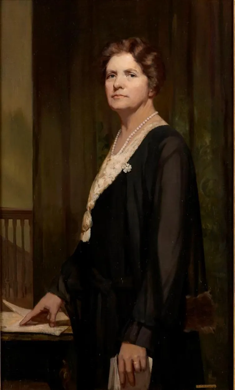 A portrait of a white woman with brown wavy hair. She is wearing a white blouse with pearls and a see through cardigan.