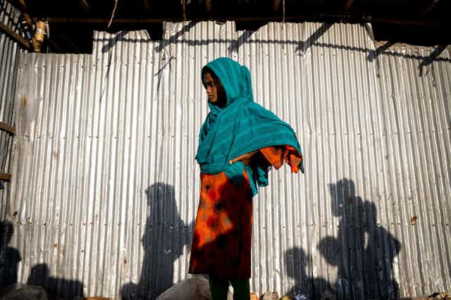 A hooded, young woman stands against a corrugated iron wall.