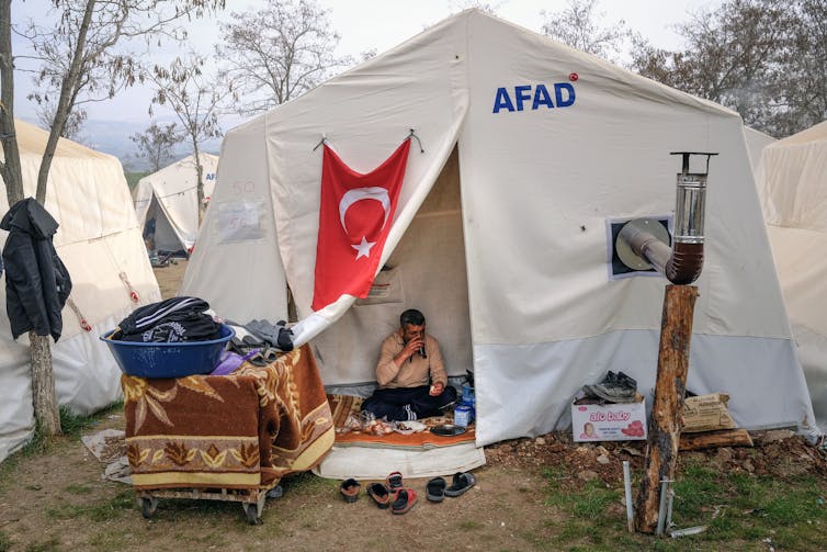 A person sits cross-legged in a white tent bearing the Turkish flag.