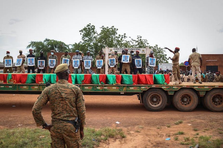Des hommes portant des uniformes militaires s'agenouillent sur des cercueils drapés de rouge et de vert qui se trouvent à l'arrière d'un camion.