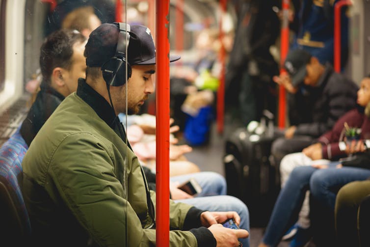 A young man in a green jacket listens to music on headphones while travelling on the London Underground.
