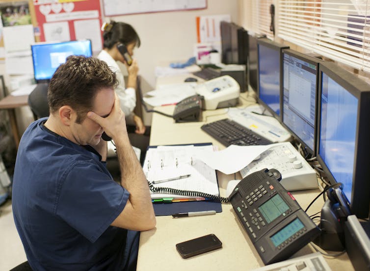 Two health care workers on the phone at computer hub, one with hand covering eyes.