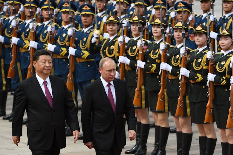 Two men in suits walk in front of a formal display of military personnel, all wearing blue and yellow outfits and carrying rifles.