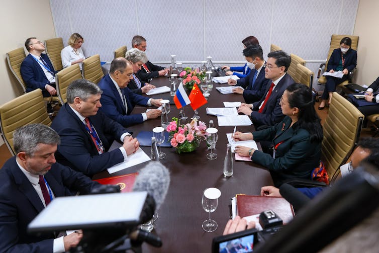 People are shown sitting around a long table that has a Russian and Chinese flags on it.