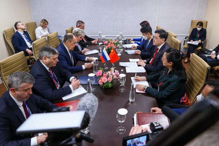 People are shown sitting around a long table that has a Russian and Chinese flag on it.