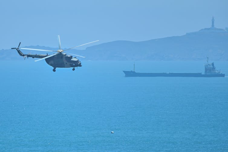 A helicopter is seen over a blue ocean, with land in the distance.