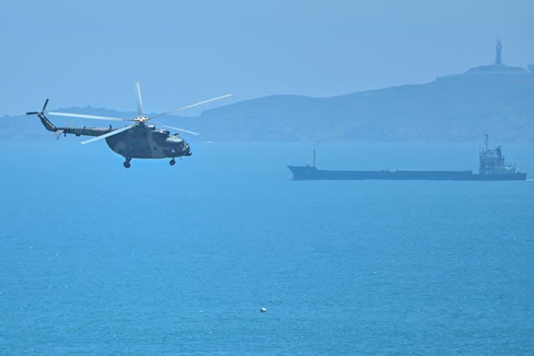 Se ve un helicóptero sobre un océano azul, con tierra a lo lejos.