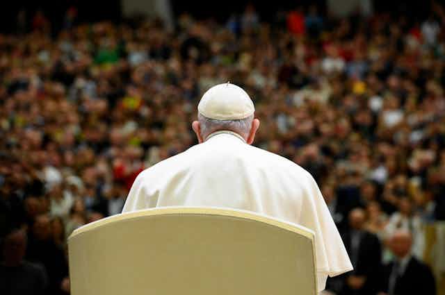 Photo Francis, wearing a white skullcap, sits facing a large crowd.
