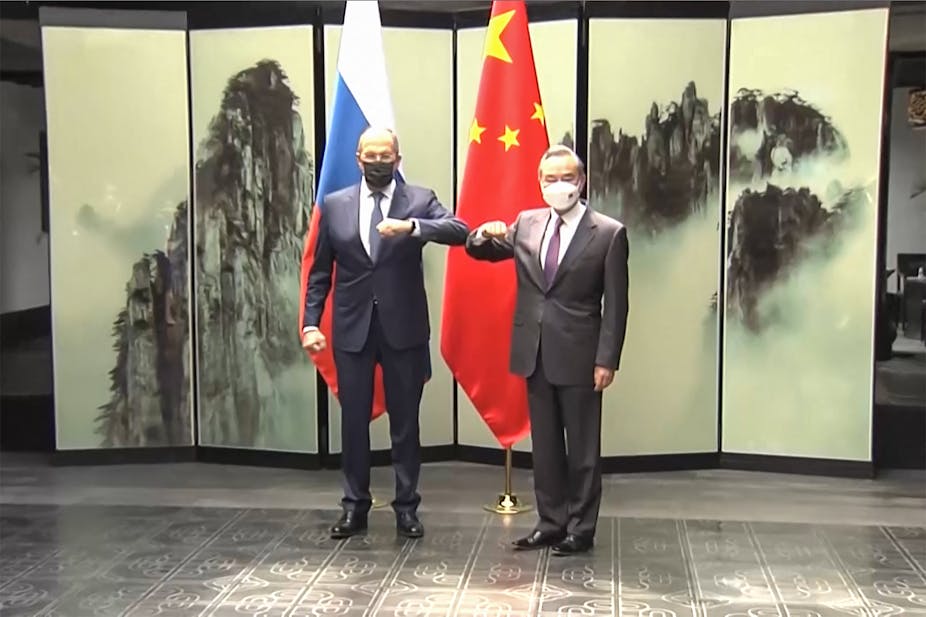 Two men wearing suits and face masks bump elbow, standing in front of Russian and Chinese flags. 