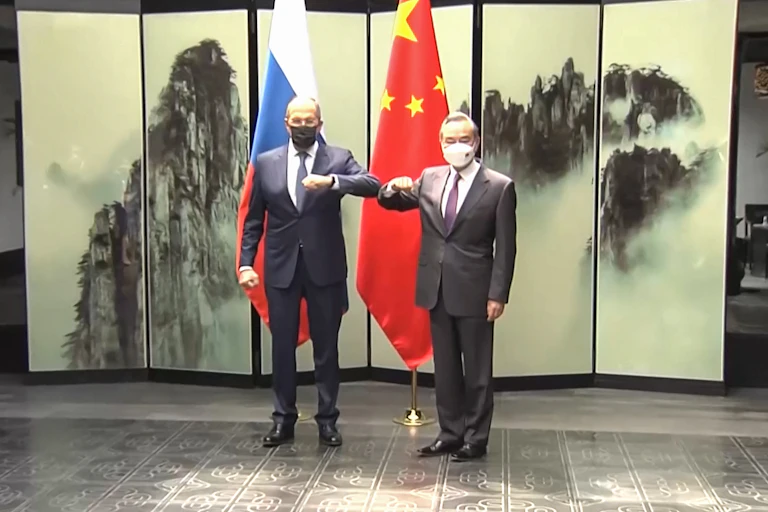Two men wearing suits and face masks bump elbow, standing in front of Russian and Chinese flags.