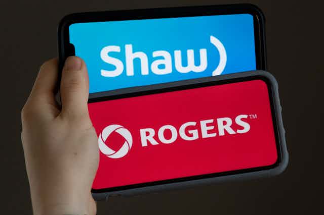 A hand holds two cellphones, one displaying the Shaw logo and one displaying the Rogers logo