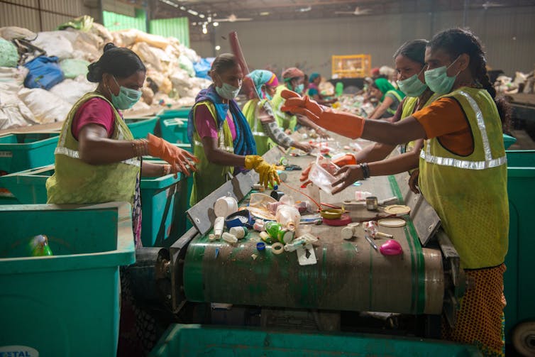 Women wearing masks and fluorescent worker vests sort through trash on a conveyor belt