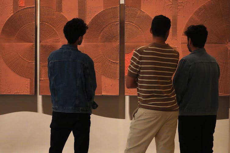Men stand looking at an artwork that is an ochre coloured painting with sculptural circles raised from its surface.
