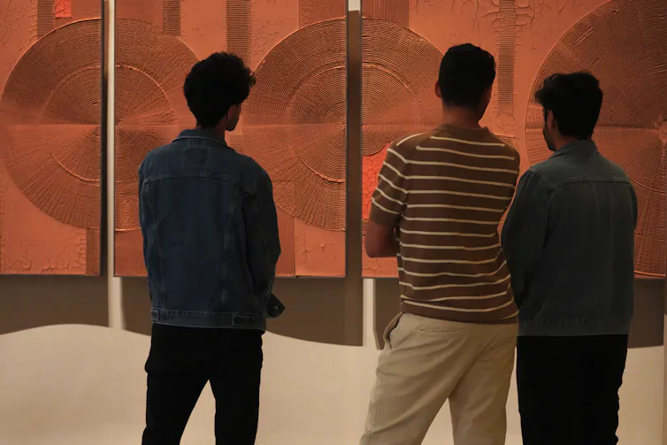 Men stand looking at an artwork that is an ochre coloured painting with sculptural circles raised from its surface.