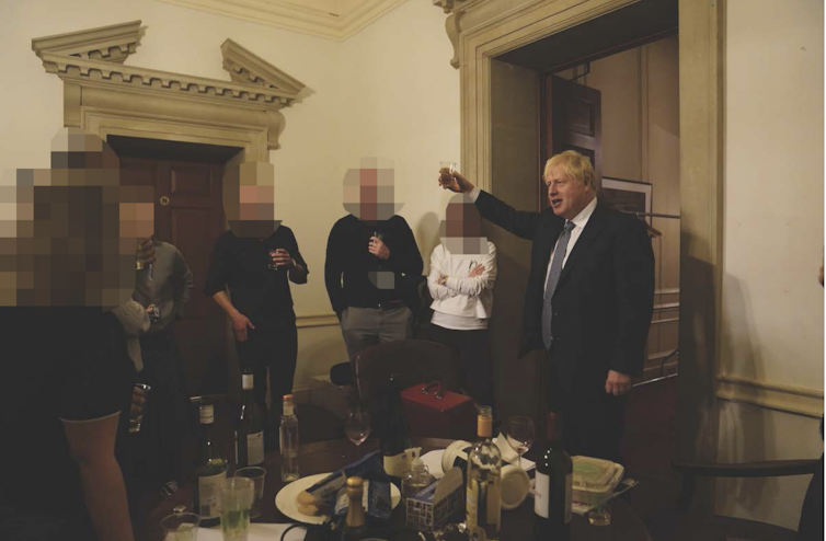 Boris Johnson raising a glass next to colleagues in front of a table covered in wine bottles.