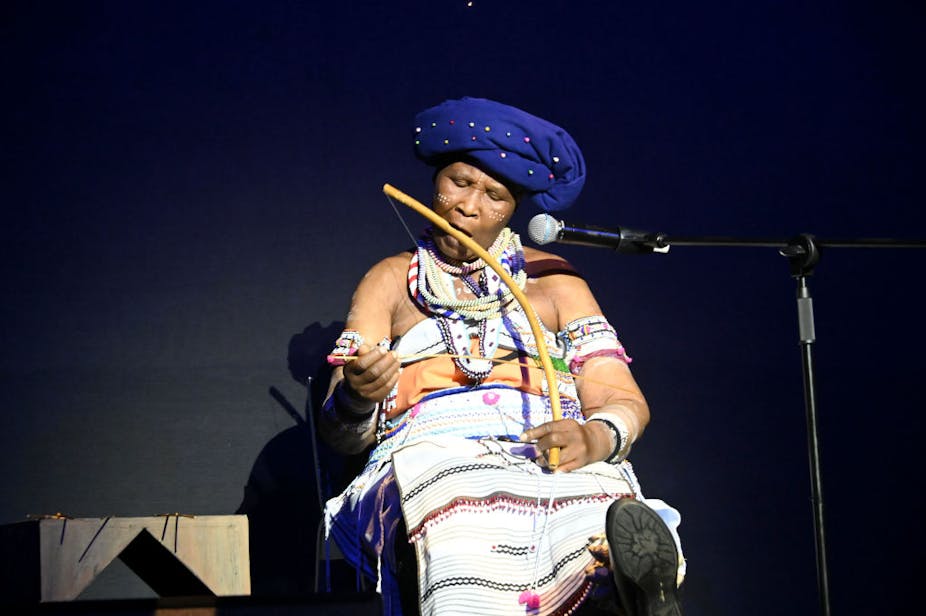 A woman sits on stage near a microphone, dressed in traditional African attire and blowing into a wooden bow which has a string on it which she plays with a stick.