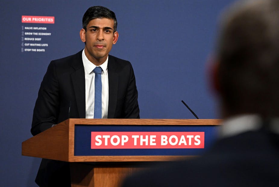 Prime Minister Rishi Sunak stands at a podium that reads 'stop the boats' on a red banner
