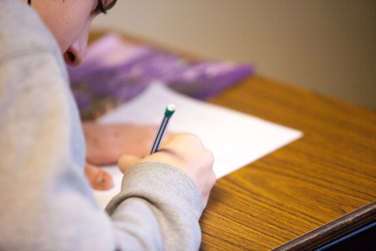 A young man is seen from behind as he writes an exam, pen in hand.