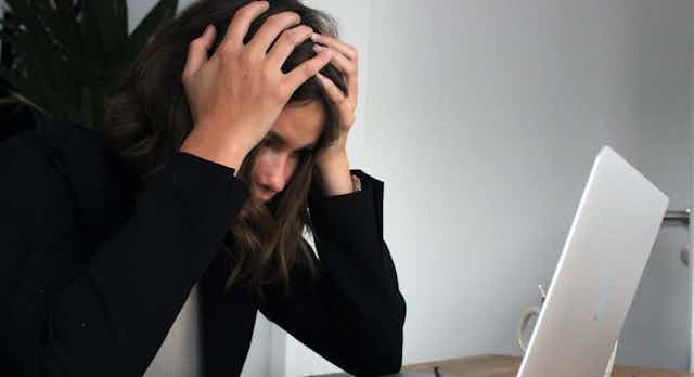 A young woman with dark hair has her hands to her head as she stares at her laptop.