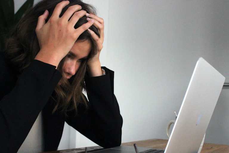 A young woman with dark hair has her hands to her head as she stares at her laptop.