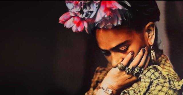 A pensive Frida Kahlo with flowers in here hair and several rings on her left hand.