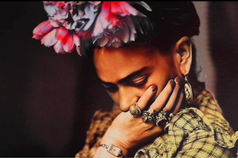 A pensive Frida Kahlo with flowers in here hair and several rings on her left hand.