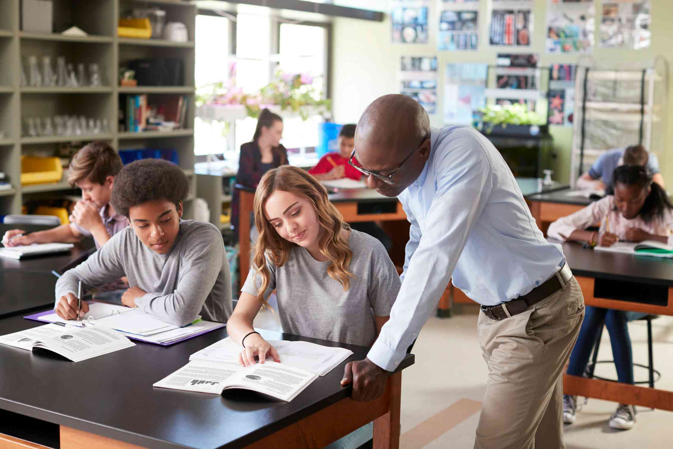A teacher leans over the desk of two students and looks at the notes of one.