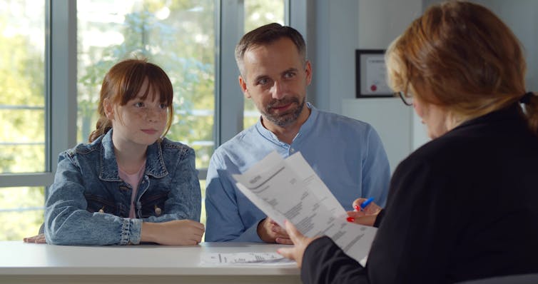 A man and his daughter, who has long brown hair and bangs, talk at a desk with a teacher, who is seen from behind.