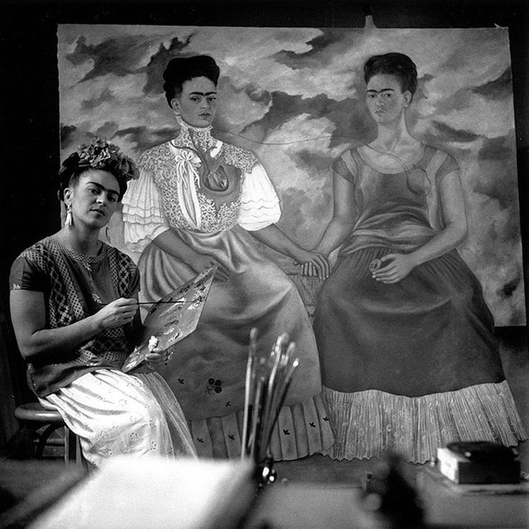 A woman called Frida Kahlo standing by a painting of two women who look like her.
