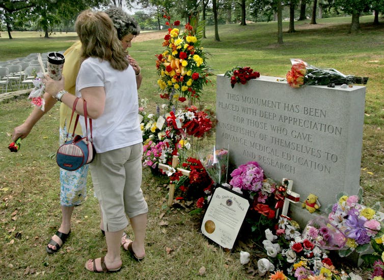 Women embrace each other at a grave strewn with flowers.